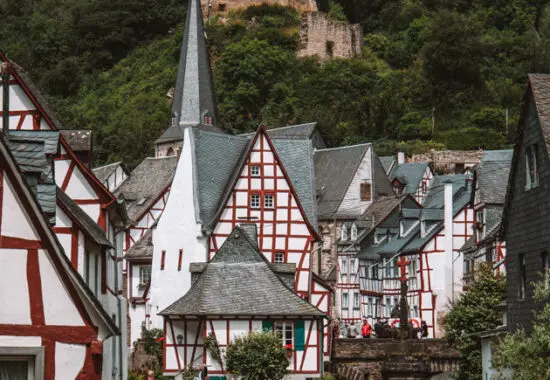 the town of monreal, germany. There is a river that runs through the middle of the town with half timbered red, white and blue houses on each side. In the background is a church with a couple getting married and the church ruin löwenburg