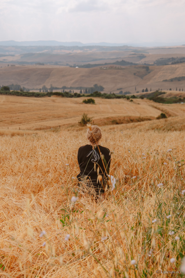 girl sat in a wheat field with rolling hills behind her