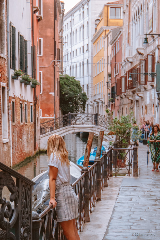 girl standing looking over one of the thin venice canals with colourful houses in the background