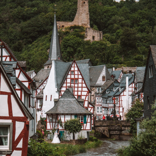 the town of monreal, germany. There is a river that runs through the middle of the town with half timbered red, white and blue houses on each side. In the background is a church with a couple getting married and the church ruin löwenburg