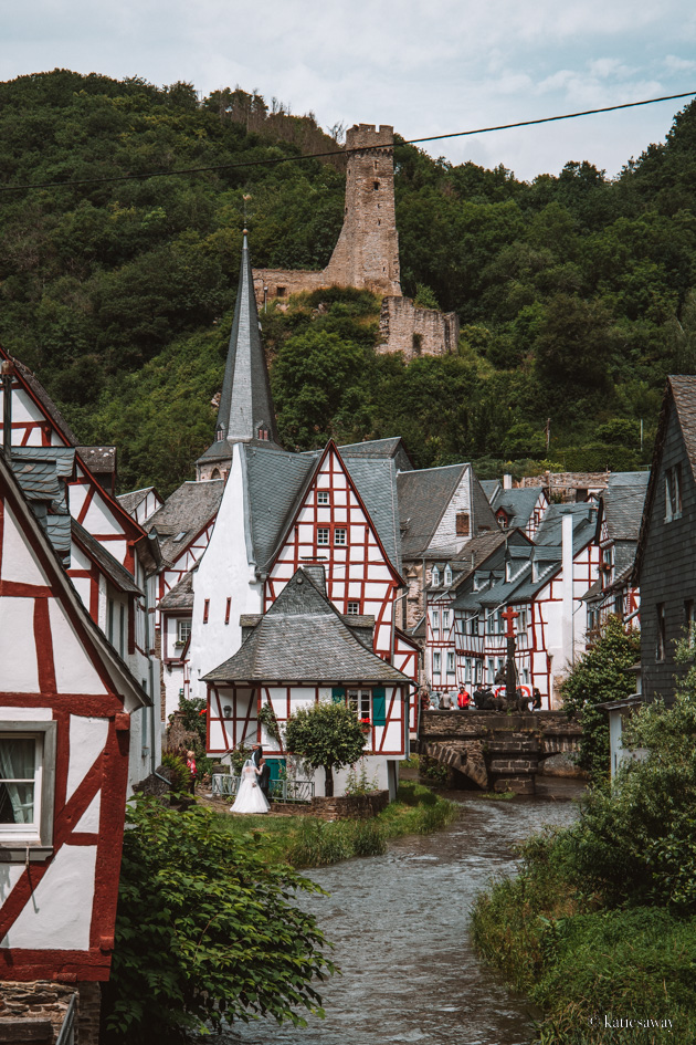 the town of monreal, germany. There is a river that runs through the middle of the town with half timbered red, white and blue houses on each side. In the background is a church with a couple getting married and the church ruin löwenburg