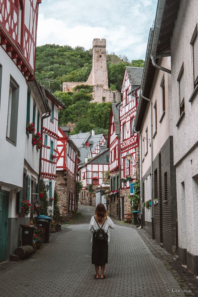 Girl in a white shirt and black trousers standing in the middle of the road in Monreal, Germany. The sides of the street are lined with red, blue and white half-timbered houses and there is löwenburg castle ruin in the background