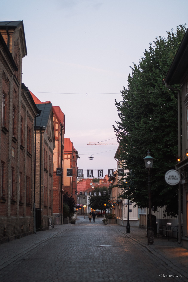 the street of haga in Gothenburg at sunset. there are tall brick buildings on each side and a black and white flag hanging across the road