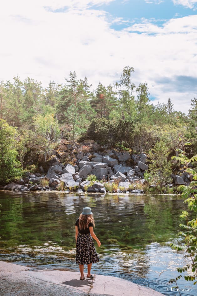 a girl in a skirt and hat walking down to the water. the water is greeny blue with big boulders on the land opposite