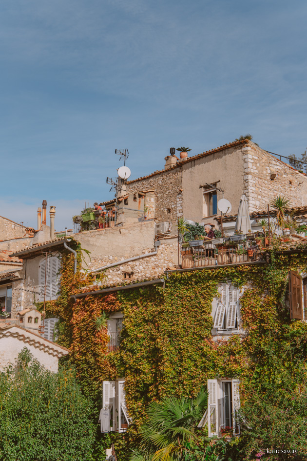 stone buildings in St Paul de Vence covered in a climbing green plant, with overlapping balconies and a lady watering her plants